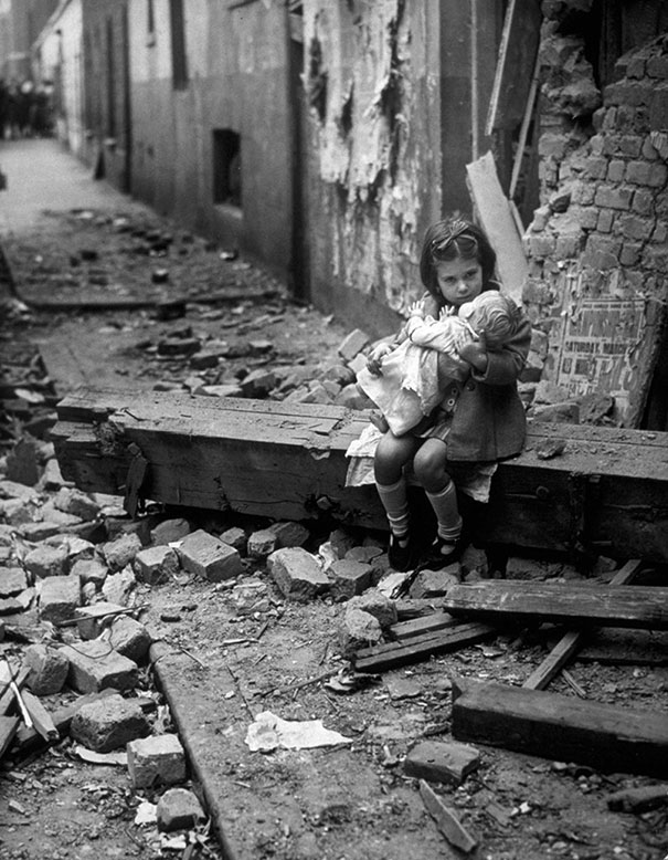 Little girl with her doll sitting in the ruins of her bombed home, London, 1940.